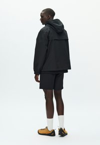 Čierna nepremokavá bunda s kapucňou, nosená cez čierne šortky. Model má na nohách žlto-oranžové turistické topánky a biele ponožky, na svetlom pozadí.