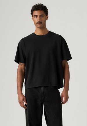 Uomo con capelli ricci e baffi che indossa una maglietta nera larga e pantaloni neri, in piedi davanti a uno sfondo chiaro e uniforme.