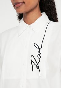 Camicia bianca con bottoni e colletto, caratterizzata da un dettaglio ricamato nero di firma e una tasca frontale. Tessuto in cotone liscio.