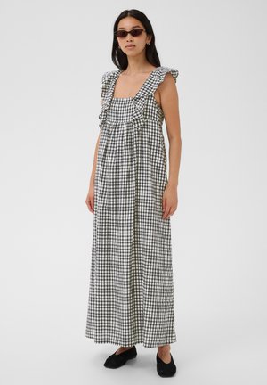 Frau trägt ein schwarz-weiß kariertes ärmelloses Maxikleid mit Rüschen, schwarze Slip-On-Schuhe und kleine ovale Sonnenbrillen vor einfarbigem Hintergrund.