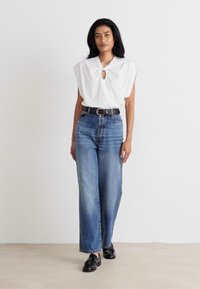 Witte, korte mouwen top met een keyhole-detail, gecombineerd met hoge taille, lichtblauwe, wijde jeans en zwarte loafers. Neutrale achtergrond.