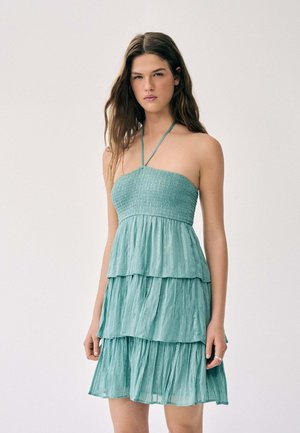 Jeune femme portant une robe sans manches à étages, turquoise clair, avec des bretelles halter, debout devant un fond clair uni.