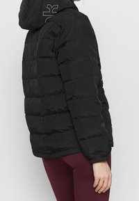 Personne portant une veste noire matelassée avec une capuche et des leggings bordeaux, debout devant un fond clair uni.