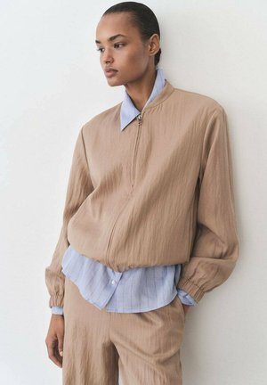 Vaalean beige vetoketjullinen bomber-takki, jossa on puffatut hihat, kerroksittain kevyen siniraidallisen paidan päällä, joka sisältää kauluksen ja tekstuuripintaa.