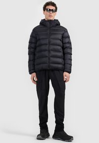 Homme portant une doudoune noire, un pantalon cargo noir, des chaussures noires et des lunettes de soleil, debout sur un fond uni gris clair.