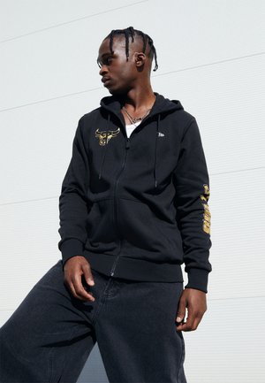 Sudadera negra con capucha y cremalleras, con un logo de toro dorado en el pecho y el texto "PUMA" a lo largo de la manga; llevada con jeans oscuros.