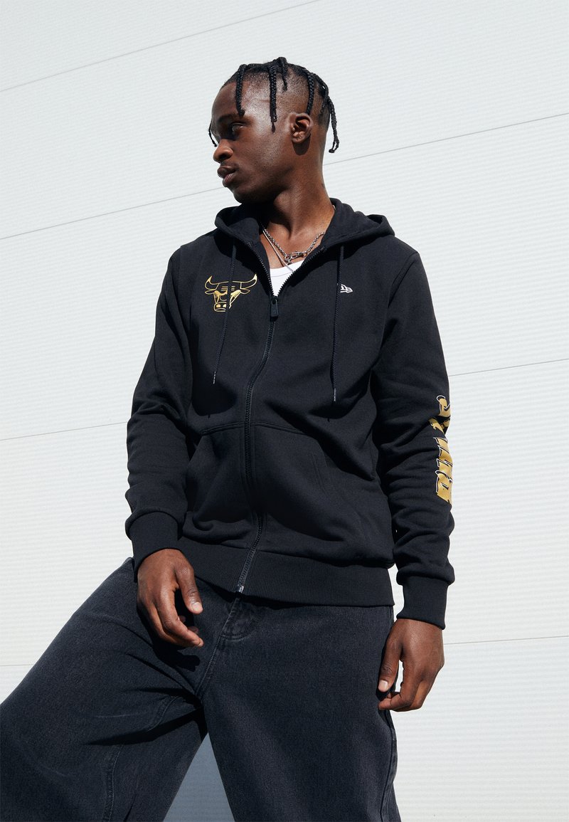 Sudadera negra con capucha y cremalleras, con un logo de toro dorado en el pecho y el texto "PUMA" a lo largo de la manga; llevada con jeans oscuros.