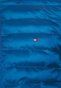 Blauwe gewatteerde pufferjack van glanzende stof met horizontale stiksels en een klein logo in rood, wit en blauw aan de voorkant.
