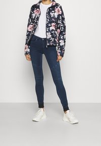 Chaqueta de plumón floral con fondo azul marino y flores blancas y rosas; jeans ajustados de mezclilla oscura con acentos acanalados; zapatillas blancas.