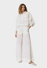 Donna che indossa una blusa bianca ampia e trasparente con dettagli floreali e pantaloni a gamba larga, abbinati a scarpe slip-on bianche con pois.
