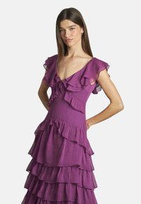 Lila schichtetes Kleid mit Rüschensäumen, V-Ausschnitt und transparenten Lagen. Der Stoff hat ein leichtes, strukturiertes Aussehen.