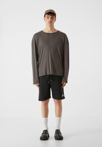 Hnedá pruhovaná košeľa s dlhým rukávom, čierne šortky s logom, biele ponožky, čierne topánky, jednofarebné pozadie. Neformálny outfit, voľný strih.