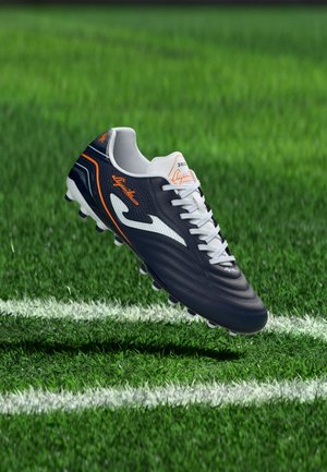 Černo-bílé kopačky s oranžovými akcenty visící nad zelenou trávou poblíž bílých čar hřiště.