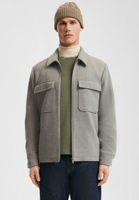 Graue Zip-Jacke mit zwei Brusttaschen, getragen über einem grünen Hemd und abgerundet mit einer beigen gerippten Strickmütze. Dunkelblaue Jeans vervollständigen den Look.