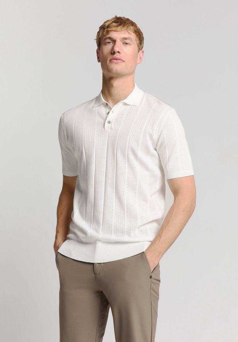 Witte polo shirt met korte mouwen, voorzien van een gestructureerd breipatroon, een drieknoopsluiting en ribgebreide rand, gecombineerd met lichtbruine broeken.