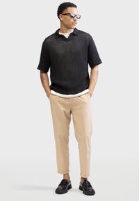 Polo en tricot noir à manches courtes, associé à un pantalon léger beige et des mocassins noirs. Doté d'un col intérieur blanc.