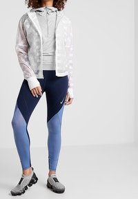Nike jacket with white semi-transparent material featuring "AIR" print, gray zip-up top, navy-blue and light blue leggings, and gray sneakers.