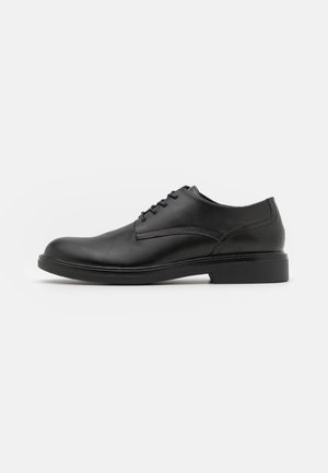 Chaussure de ville en cuir noir avec fermeture à lacets, bout rond, détails de couture minimalistes et semelle en caoutchouc plate. Vue latérale.