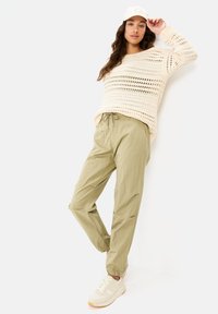 Cremfarbener Häkelpullover mit weitem Strick, kombiniert mit hellolivenen Jogginghosen mit Kordelzug und beigen Sneakers. Das Outfit hat eine lässige, lockere Passform.