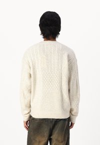 Krämfärgad stickad tröja med strukturerade kabelmönster. Har långa ärmar och en rund halsringning, i kombination med urtvättade denimjeans.