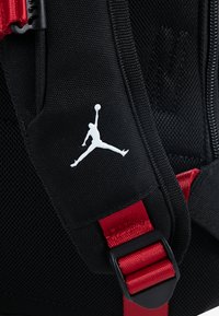 Fekete hátizsák, amelyen kiemelkedő fehér Jumpman logó található, piros állítható pántokkal és texturált hálós részletekkel a légáteresztőség érdekében.