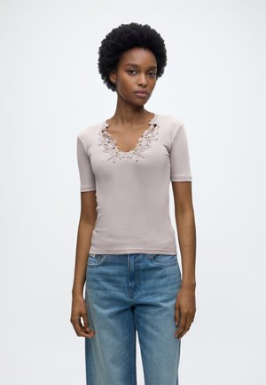 Kvinde med kort krøllet hår iført en lyserød kortærmet top med blonder ved halsudskæringen og blå højtaljede jeans, stående mod en ensfarvet baggrund.