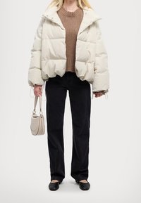 Femme portant une doudoune écrue, un pull en maille marron, un pantalon noir, des ballerines noires et tenant un sac à main beige sur un fond blanc.