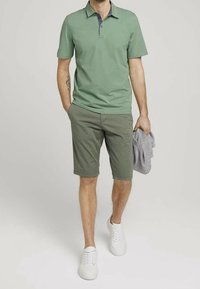 Polo-shirt en coton vert clair à manches courtes et col marine, associé à un short vert olive et des baskets blanches, tenant un pull gris.