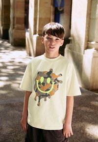 Light yellow t-shirt with a camouflage smiley graphic in orange and green. Short sleeves, relaxed fit. Worn with dark shorts.