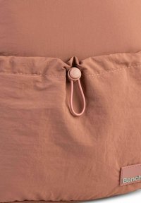 Weiche, staubige rosa Stofftasche mit gerafft oben, ausgestattet mit einem zylindrischen Toggle-Verschluss und einem strukturierten Etikett in der Ecke.