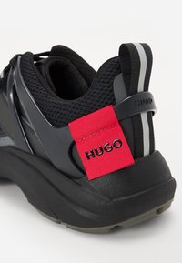 Baskets de sport noires avec une tige en mesh agrémentée d'accents brillants, une étiquette rouge portant la mention "HUGO", et une semelle extérieure noire texturée.