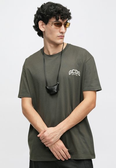 Olivnozelena majica s kratkimi rokavi in logotipom "GCDS", v kombinaciji z sončnimi očali rjave barve in malo črno torbico, ki visi na traku.