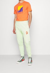 Muž v oranžovém tričku Nike, světle zelených teplákách Nike se třemi logy a zelených vysokých teniskách Nike s červenými "swoosh".