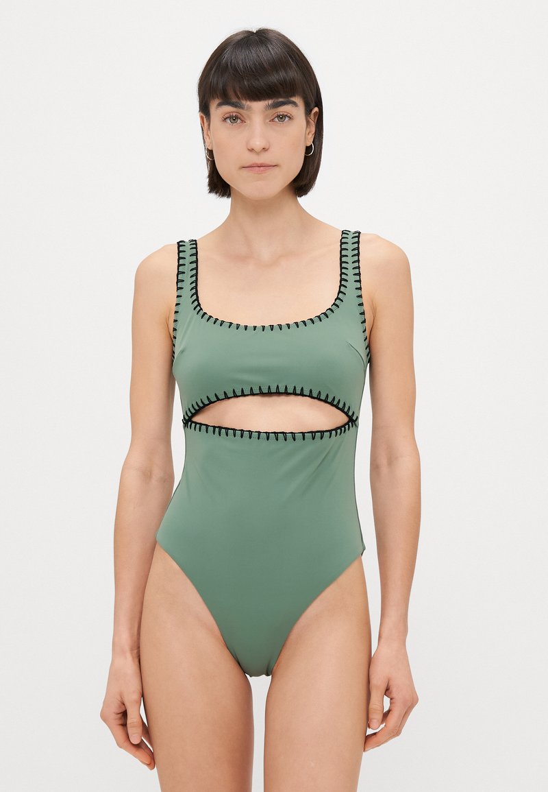 Zaļa viengabala peldkostīms ar kvadrātu dekoltē un sānu izgriezumiem, ar melnu kontrastējošu šuvju apdari. Gluda, elastīga auduma.