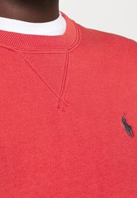 Felpa rossa con collo a coste e piccolo logo ricamato in nero di un cavallo e cavaliere sul petto, indossata sopra una camicia bianca.