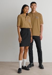 Nainen ja mies seisovat; molemmat pukeutuneet ruskeisiin polo-paitoihin, joissa on logot, mustiin alaosiin ja mustiin kenkiin; naisella on jalassaan valkoiset sukat ja hame.