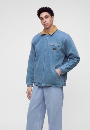 Jeune homme portant une veste en jean bleu clair avec un col marron et un jean bleu clair, debout devant un fond clair uni.