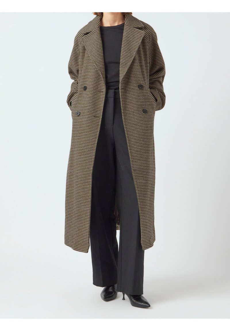 Abrigo largo de patrón houndstooth en tonos marrón y negro, de corte holgado, con doble botonadura en la parte delantera y bolsillos laterales, llevado sobre pantalones negros.