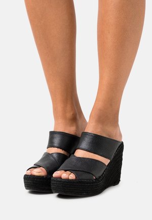 Feet wearing black leather wedge sandals with wide straps and textured platform soles on a white background.