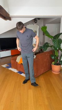 Mees, kes kannab halli polot ja pükse, reguleerib oma särki elutoas, kus on pruun diivan, toataim, teler ja puitpõrand.