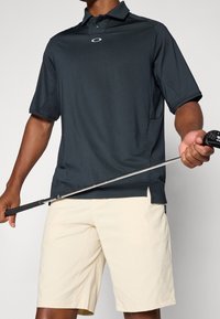 Polo shirt noir à manches courtes avec col, arborant un logo sur la poitrine. Shorts beige clair. Personne tenant un club de golf. Texture de tissu lisse.