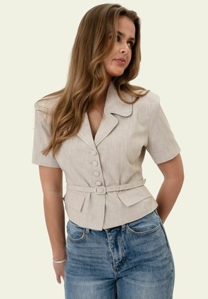 Frau mit langen braunen Haaren, die einen beigen, kurzärmeligen Blazer mit Knöpfen und Gürtel sowie blaue Jeans trägt, schaut nach links.