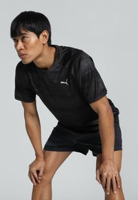 Sort atletisk T-shirt med et subtilt strukturmønster, der har et hvidt Puma-logo på brystet. Parret med sorte shorts, begge dele er lette.