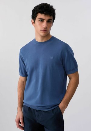 Giovane uomo con capelli neri ricci, che indossa una maglia blu a maniche corte e pantaloni scuri, in piedi davanti a uno sfondo chiaro e uniforme.