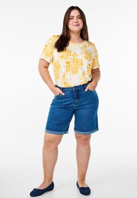 Lühikeste varrukatega kollane lillemuster top, sinised denim lühikesed püksid keerdküpsistega ja tumesinised korterid. Mudel seisab käed taskutes valge tausta against.