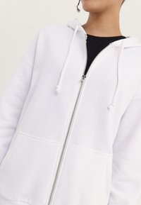 Felpa bianca con zip e cappuccio regolabile, caratterizzata da una texture morbida, tasche frontali e dettagli della cerniera in argento.