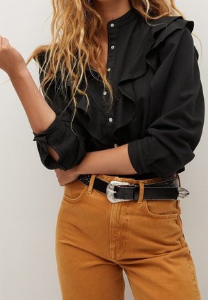 Femme portant un chemisier noir à volants boutonné rentré dans un pantalon moutarde taille haute avec une ceinture noire à boucle argentée.