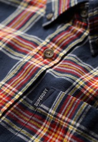 Chemise à carreaux bleu marine, rouge, jaune et blanc boutonnée avec une petite étiquette noire « Superdry » sur la poche.