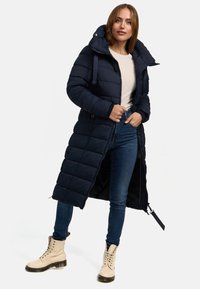 Navy puffer coat with a hood, zip closure, and a long silhouette. Worn over a light top and paired with blue jeans and beige lace-up boots.