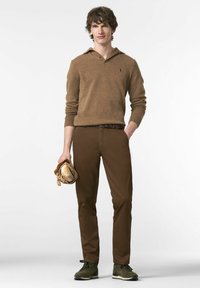 Pull en tricot marron avec un col, associé à un pantalon marron ajusté. Le mannequin tient une miche de pain enveloppée dans du papier. Des baskets olive complètent le look.